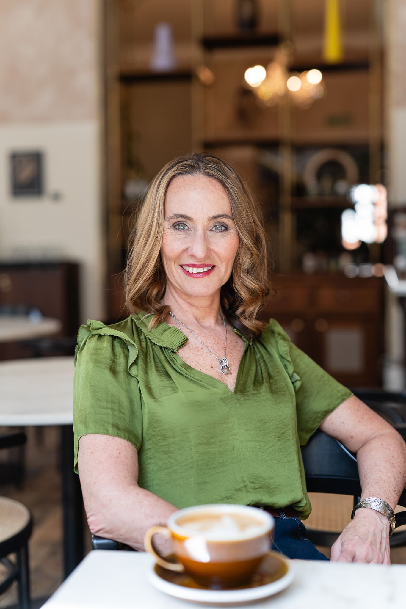 woman in a green top sitting at desk with coffee, looking straight to the camera